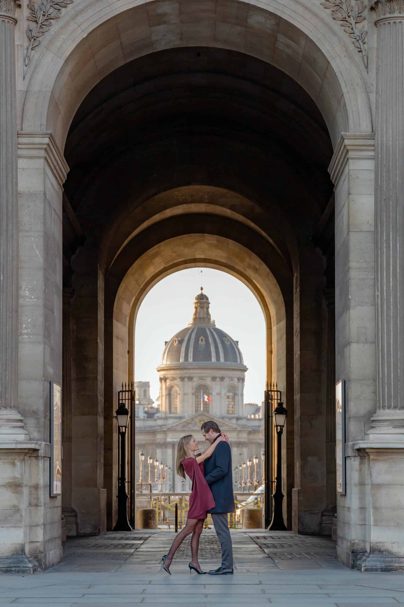 Couple embracing under the Louvre archway with the Institut de France dome at sunset, Paris